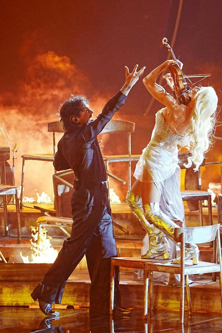 Eine Geigerin in silbernen Stiefeln spielt auf einem Stuhl, während ein Tänzer seine Hand ausstreckt.