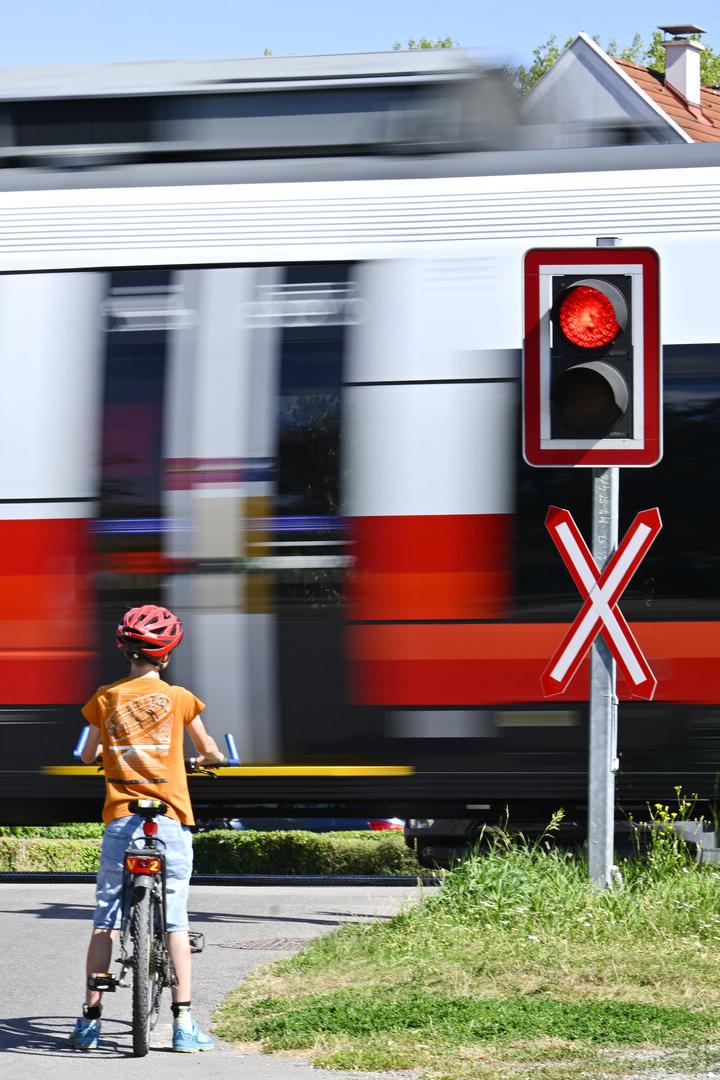 Ein Kind mit Fahrrad wartet an einem Bahnübergang mit rotem Signal, während ein Zug vorbeifährt.