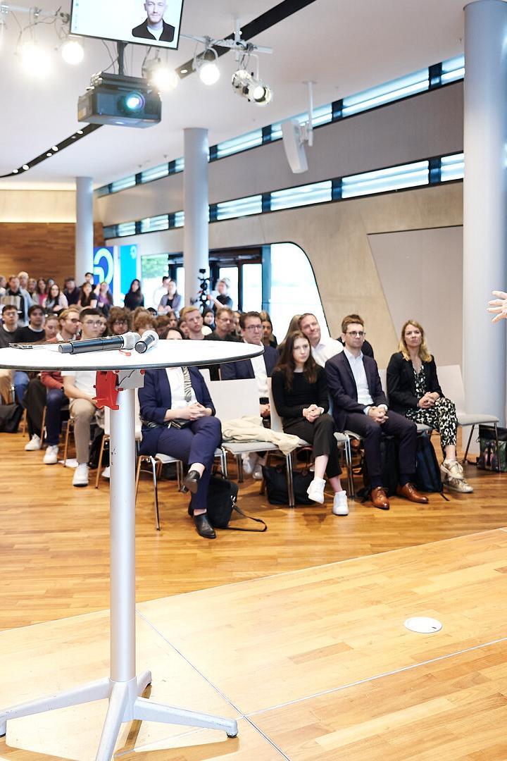 Ein Mann hält einen Vortrag auf einer Bühne vor einem aufmerksam zuhörenden Publikum in einem modernen Saal.