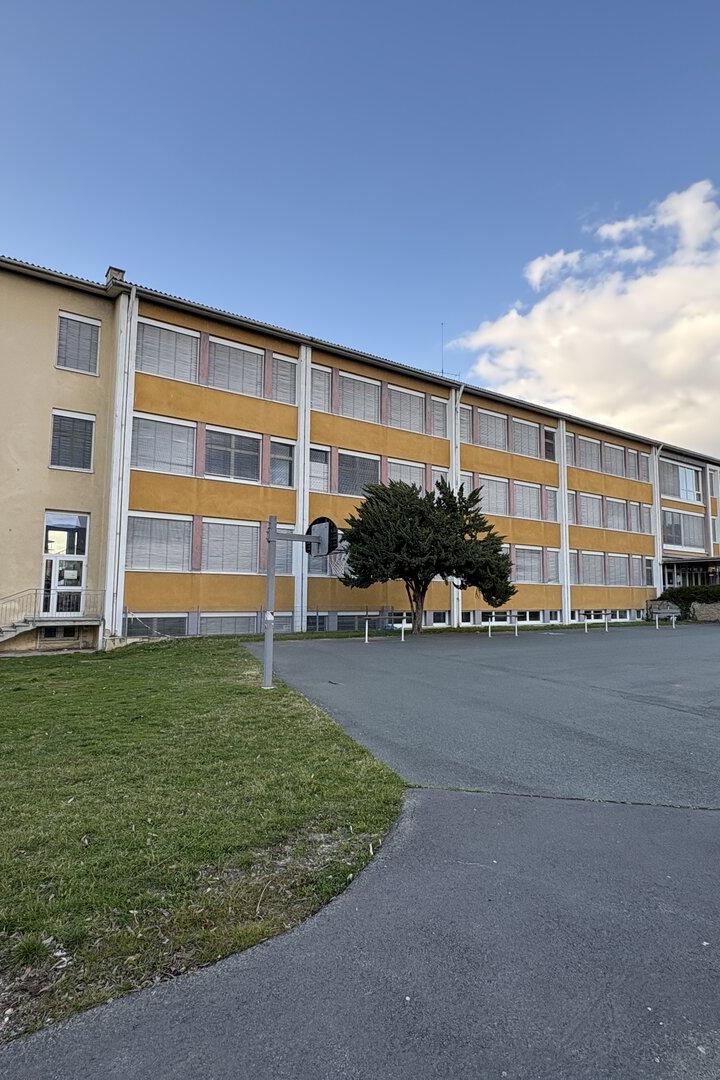 Ein langes, gelbliches Schulgebäude mit vielen Fenstern steht neben einem leeren Schulhof mit etwas Grünfläche und einem Baum.