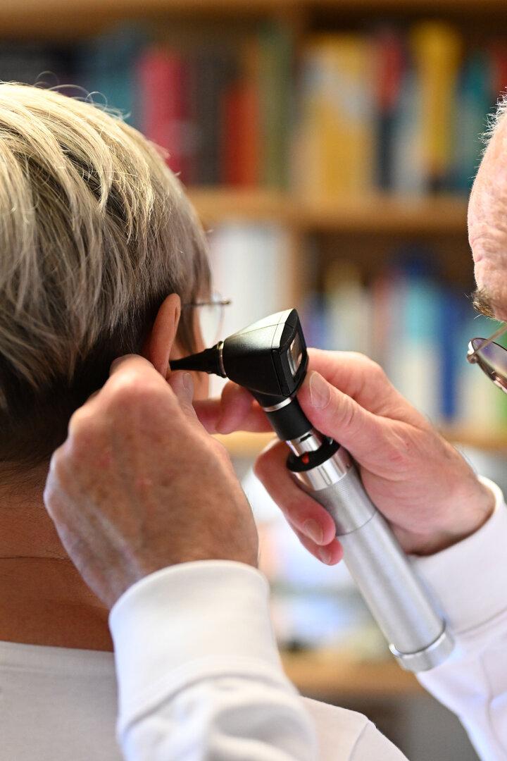 Ein Arzt untersucht mit einem Otoskop das Ohr einer Person mit kurzem, blondem Haar.