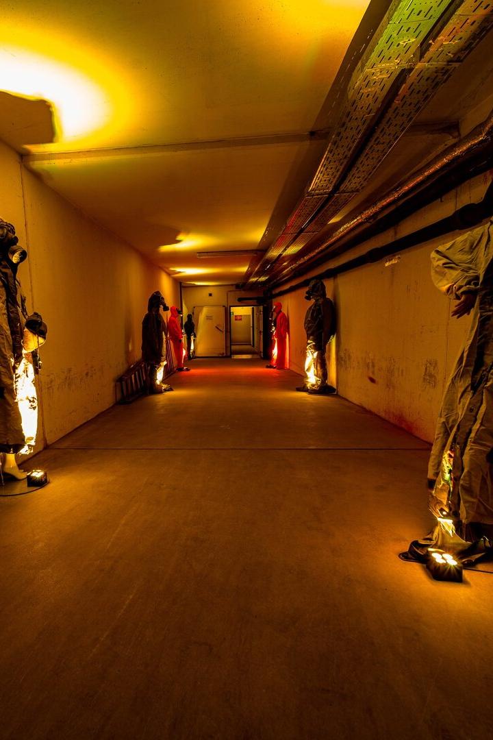 Ein langer, gelb beleuchteter Gang mit Schaufensterpuppen in Schutzanzügen und Gasmasken an den Wänden.