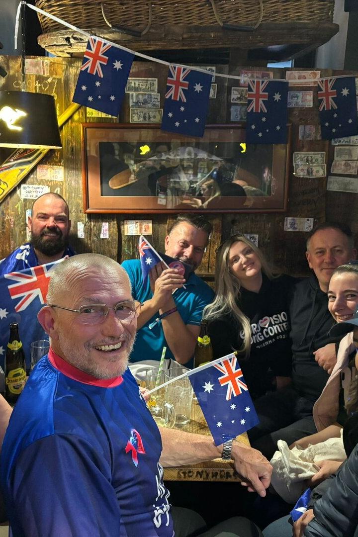 Eine fröhliche Gruppe von Menschen sitzt in einem dekorierten Raum mit vielen australischen Flaggen und lächelt in die Kamera.