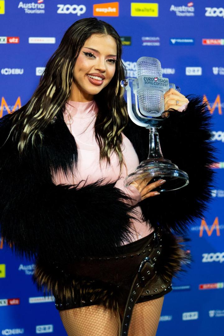 Eine junge Frau hält einen gläsernen Eurovision Song Contest Pokal und lächelt vor einer Sponsorenwand.