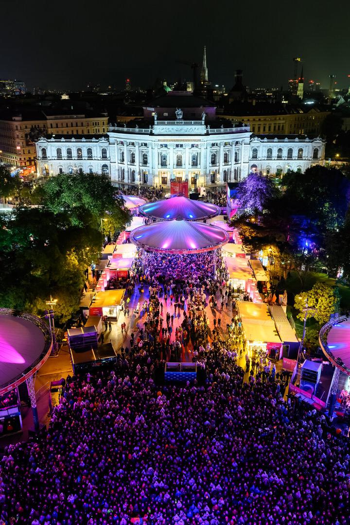 Nächtliche Menschenmenge vor der beleuchteten Wiener Burgtheater unter farbigen Zeltdächern.