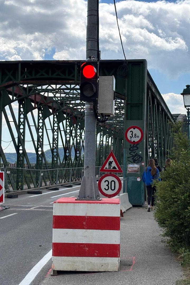Eine grüne Stahlbrücke mit Baustellenabsperrungen, Verkehrsschildern und roter Ampel, zwei Personen gehen auf dem Gehweg.