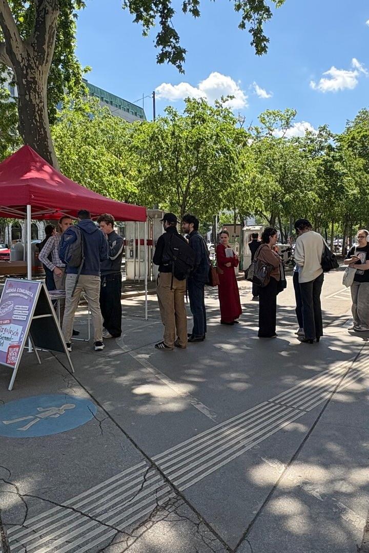 Mehrere Menschen stehen bei sonnigem Wetter an einem roten Pavillon auf einem Gehweg, einige warten in einer Schlange.