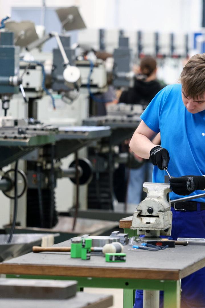 Ein junger Mann arbeitet konzentriert an einem Schraubstock in einer Werkstatt mit mehreren Maschinen im Hintergrund.