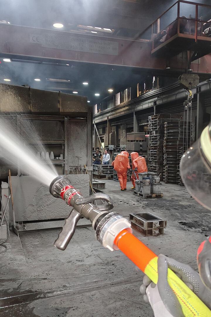 Ein Feuerwehrmann spritzt Wasser in einer Industriehalle, im Hintergrund arbeiten Personen in orangefarbenen Schutzanzügen.