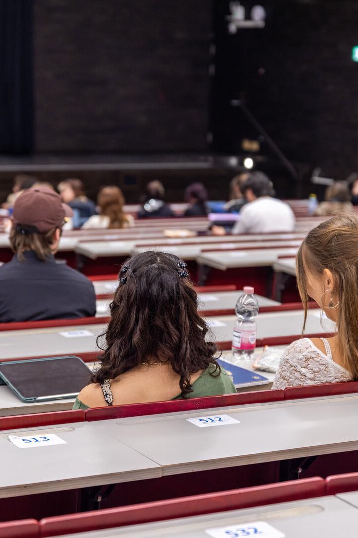 Mehrere Studierende sitzen in einem Hörsaal mit nummerierten Plätzen und schauen zur Bühne nach vorne.