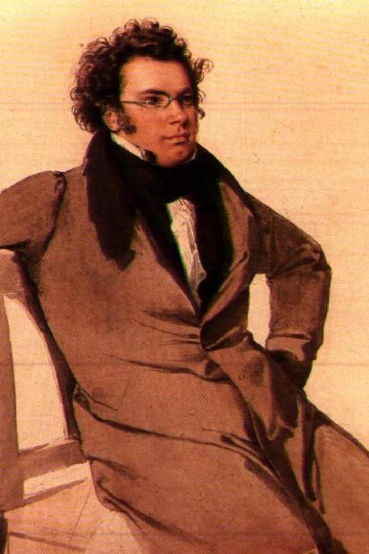 Franz Schubert sitzt lässig auf einem Stuhl, mit Brille und dunklem Mantel.