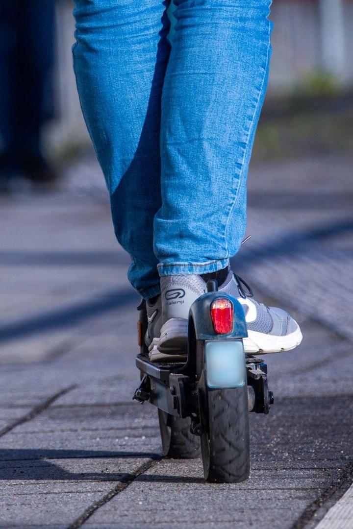 Person in blauer Jeans fährt auf einem E-Scooter auf einem gepflasterten Gehweg.