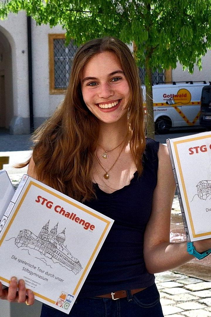 Junge Frau mit langen Haaren hält draußen mehrere Spielekartons mit der Aufschrift „STG Challenge“ und lächelt.