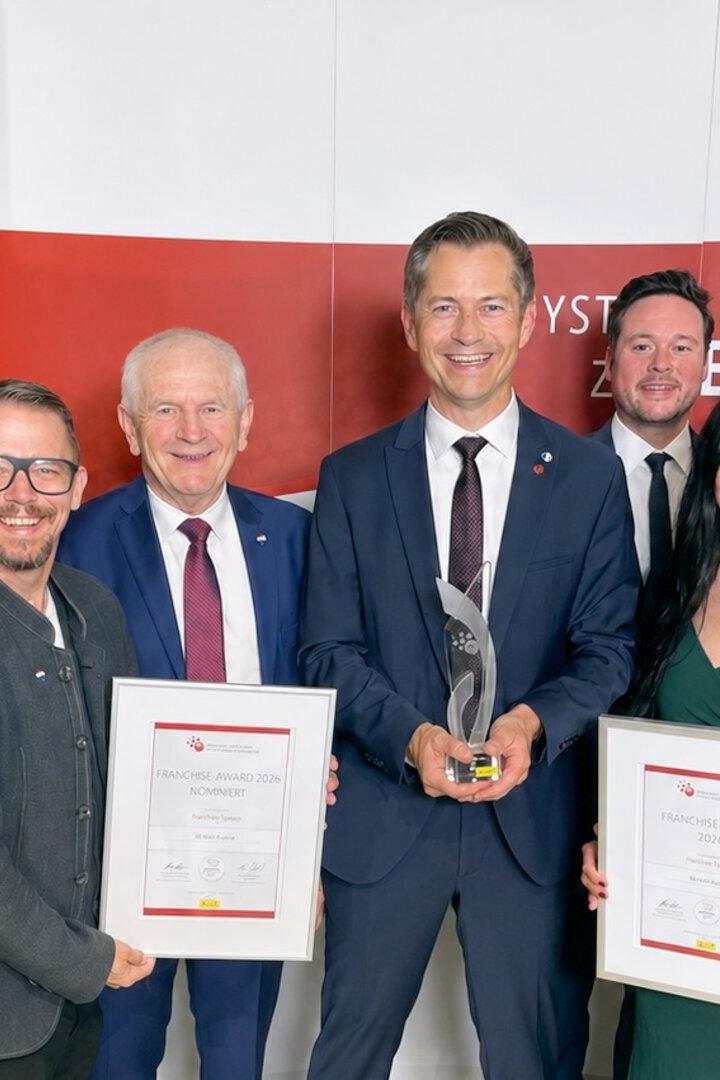 Neun Personen in festlicher Kleidung halten Urkunden und eine Trophäe vor einer Wand mit dem Logo des Österreichischen Franchise-Verbands.