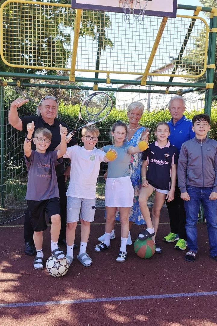 Sechs Kinder und drei Erwachsene stehen fröhlich auf einem Sportplatz vor einem Basketballkorb, einige halten Bälle in der Hand.