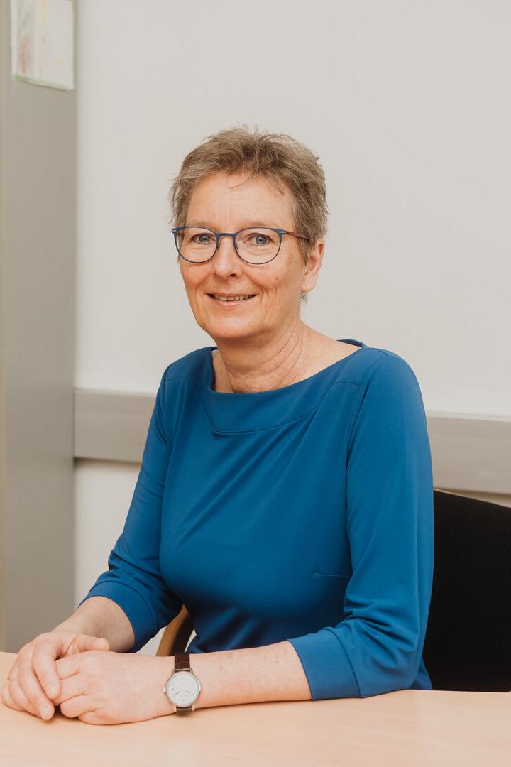 Eine Frau mit Brille und blauem Oberteil sitzt lächelnd an einem Schreibtisch in einem Büro mit Regal im Hintergrund.