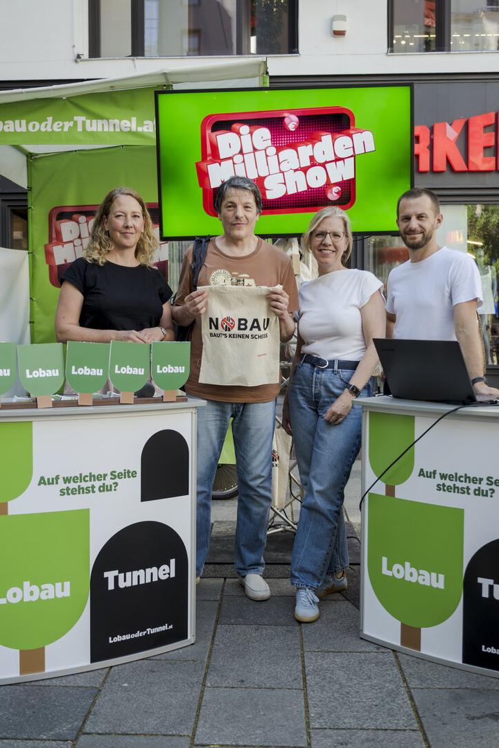 Vier Personen stehen an einem Stand mit der Aufschrift "Lobau Tunnel" und "Die Milliarden Show".