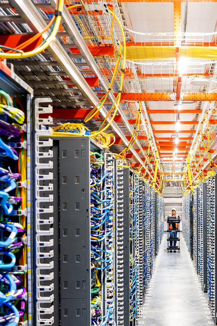 Reihen von Serverracks mit bunten Kabeln und einer Person, die einen Wagen durch einen Gang schiebt.