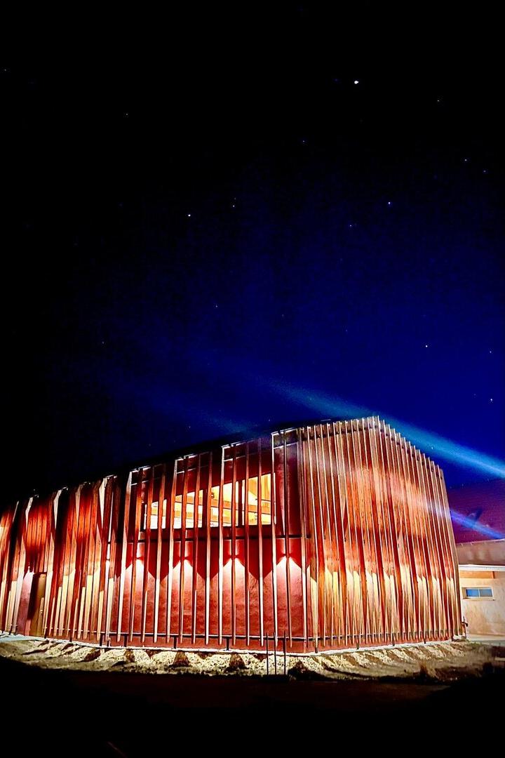 Modernes Gebäude mit vertikalen Holzlamellen, nachts beleuchtet, darüber ein klarer Sternenhimmel.
