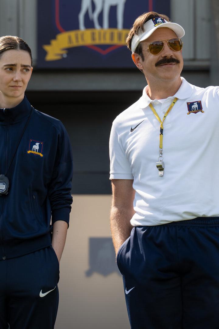 Ted Lasso zeigt auf dem Fußballfeld mit einer Frau im Trainingsanzug neben ihm.