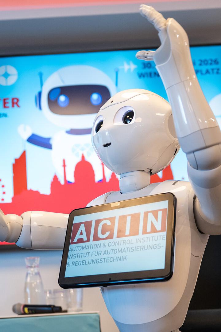 Ein humanoider Roboter mit Tablet hebt die Arme, im Hintergrund eine bunte Tafel mit dem Schriftzug „Festival der Roboter“.