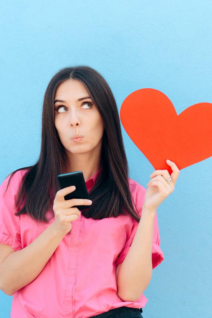 Frau in rosa Hemd hält ein rotes Herz und ein Smartphone vor blauem Hintergrund und schaut nachdenklich nach oben.