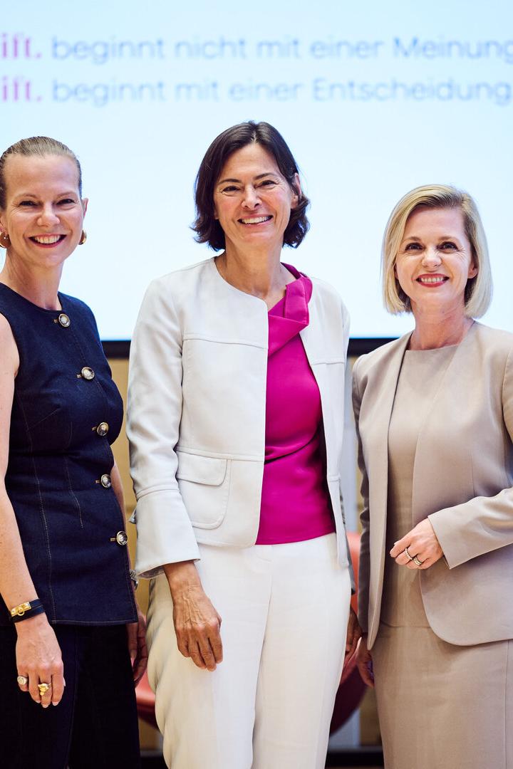 Drei Frauen stehen lächelnd nebeneinander in einem modernen Konferenzraum mit Stühlen und Präsentationsbildschirm.