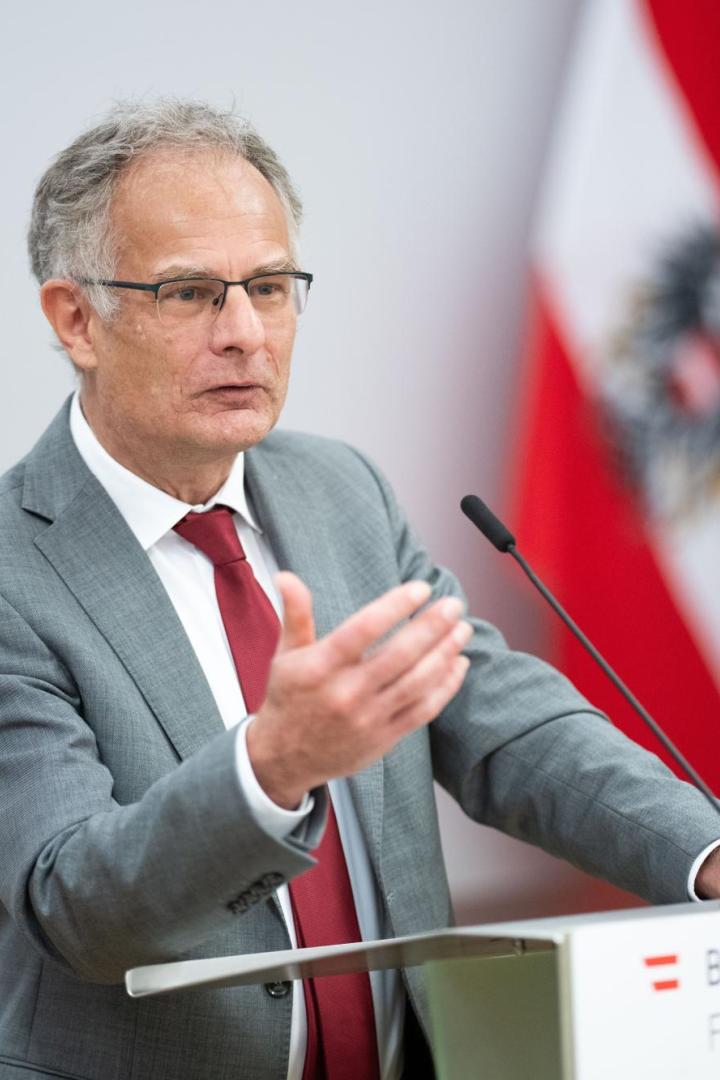 Ein Mann im grauen Anzug spricht an einem Rednerpult mit Wasserglas, im Hintergrund österreichische und EU-Flagge.
