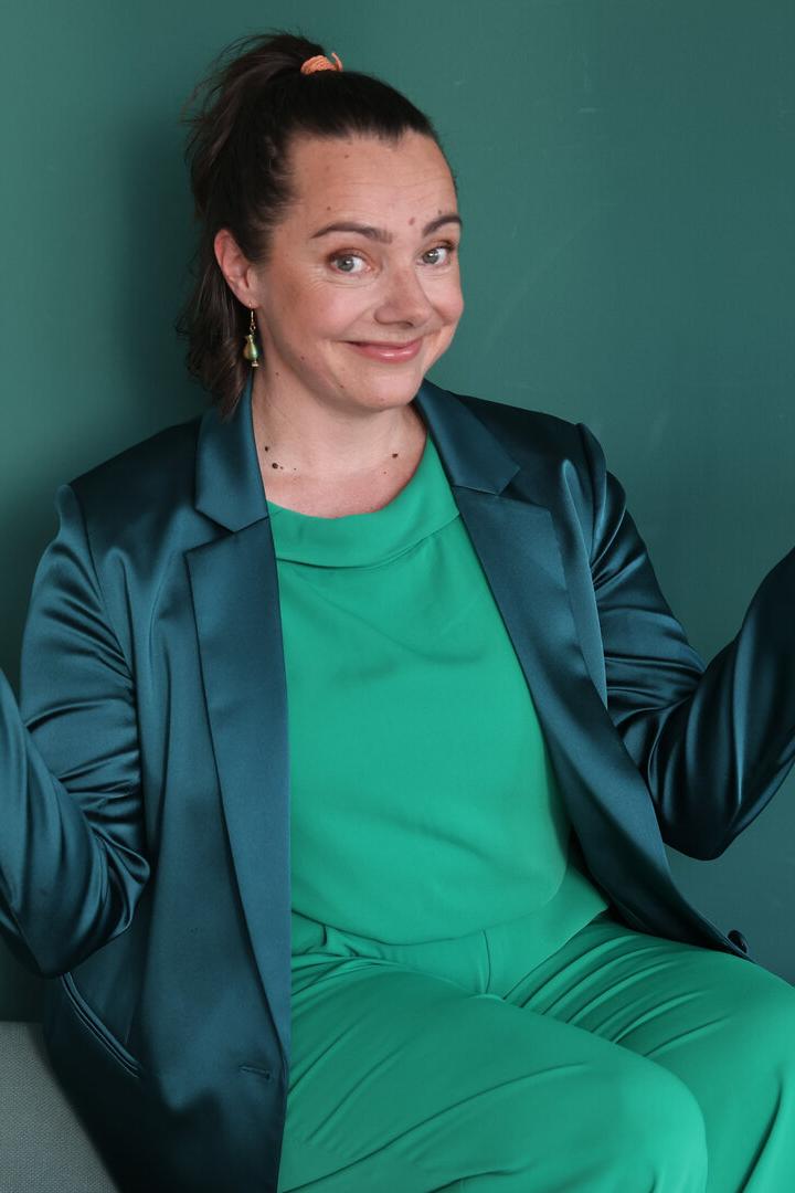 Eine Frau in grüner Kleidung und einem bläulichen Blazer sitzt mit fragenden, offenen Händen vor einem grünen Hintergrund.