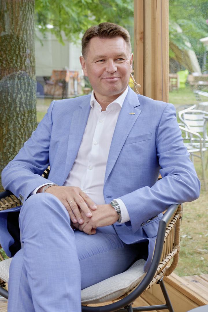 Ein Mann im hellblauen Anzug sitzt entspannt in einem Sessel vor einer Glaswand.