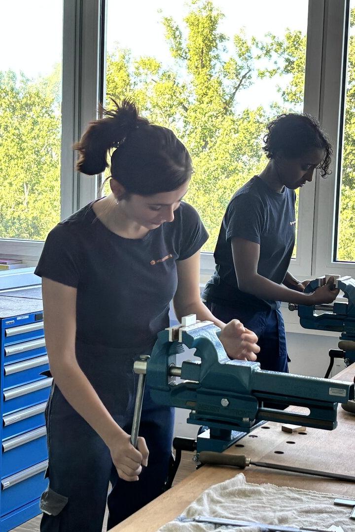 Zwei Personen arbeiten an Werkbänken mit Schraubstöcken und Werkzeugen vor großen Fenstern mit Blick ins Grüne.