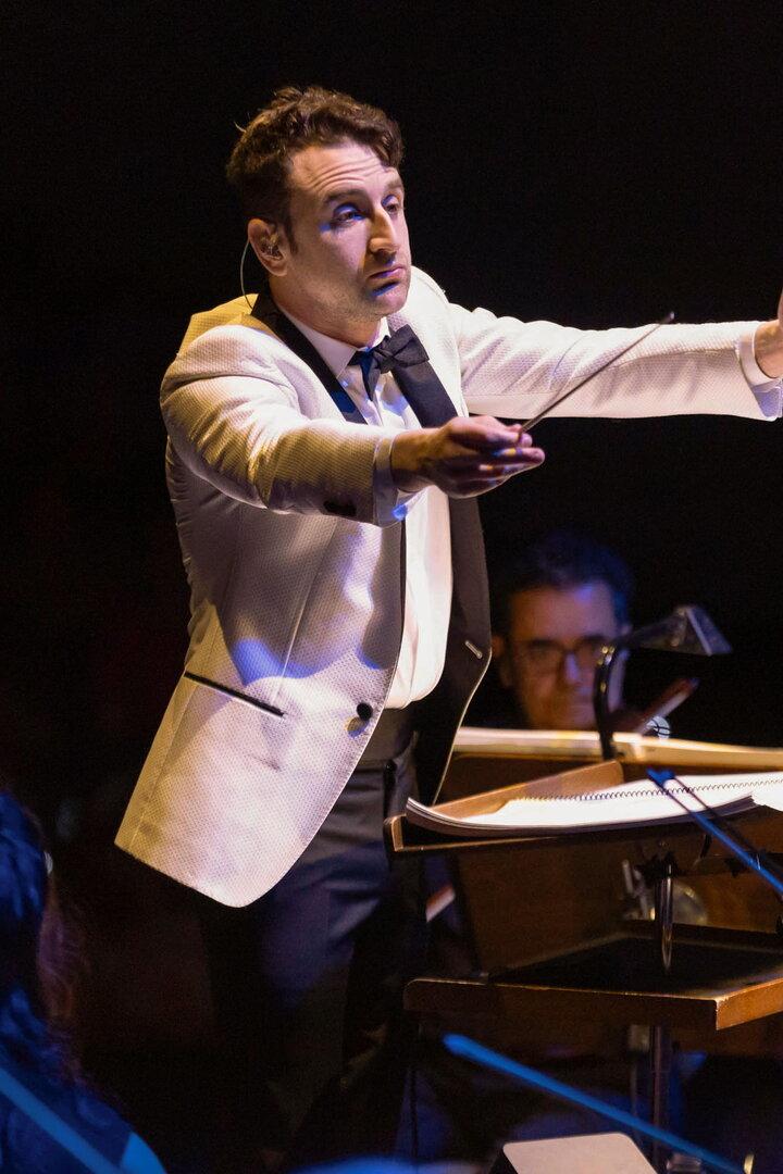 Ein Dirigent in weißem Sakko leitet ein Orchester, das konzentriert auf seine Anweisungen achtet.
