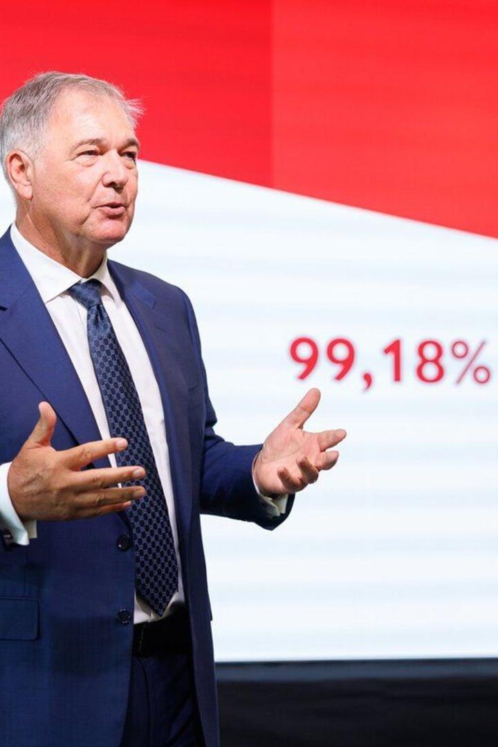 Walter Ruck  im blauen Anzug  spricht nach seiner Leinwand vor einer Leinwand, auf der  99,18 % gezeigt werden. 