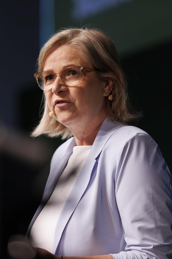 Eine Frau mit Brille und hellem Blazer spricht vor dunklem Hintergrund.