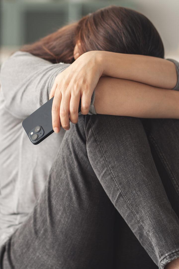 Eine Frau sitzt mit gesenktem Kopf auf einem Sofa und hält ein Smartphone in der Hand.