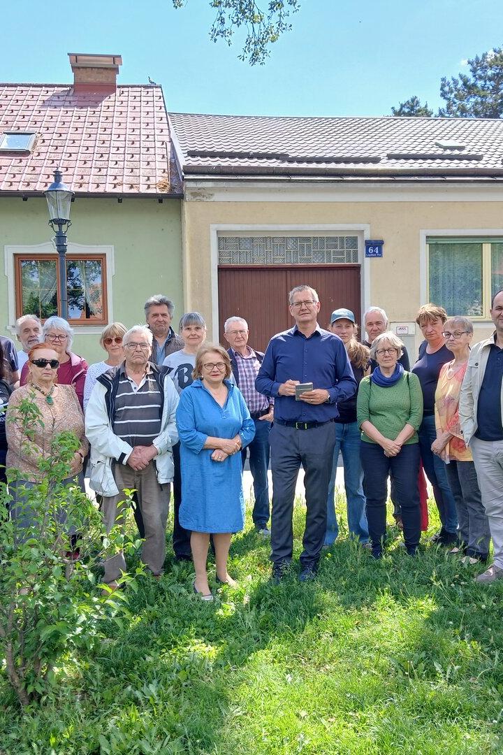 Eine große Gruppe von Menschen steht vor zwei Häusern auf einer Wiese, wobei eine ältere Frau rechts einen Rollator nutzt.