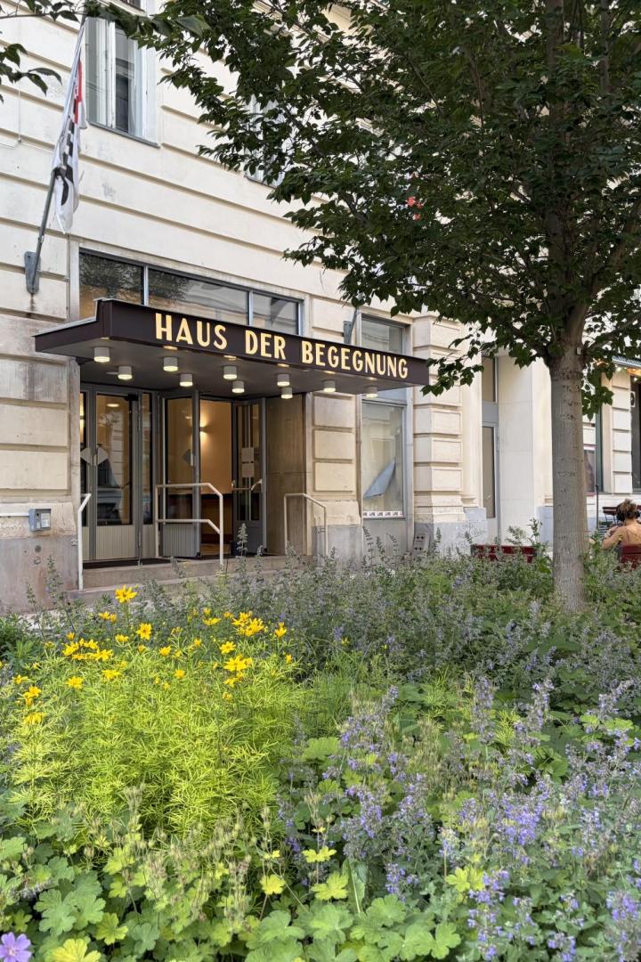Vor einem Gebäude mit dem Schriftzug „Haus der Begegnung“ wachsen Blumen und Sträucher, daneben sitzen Menschen im Straßencafé.