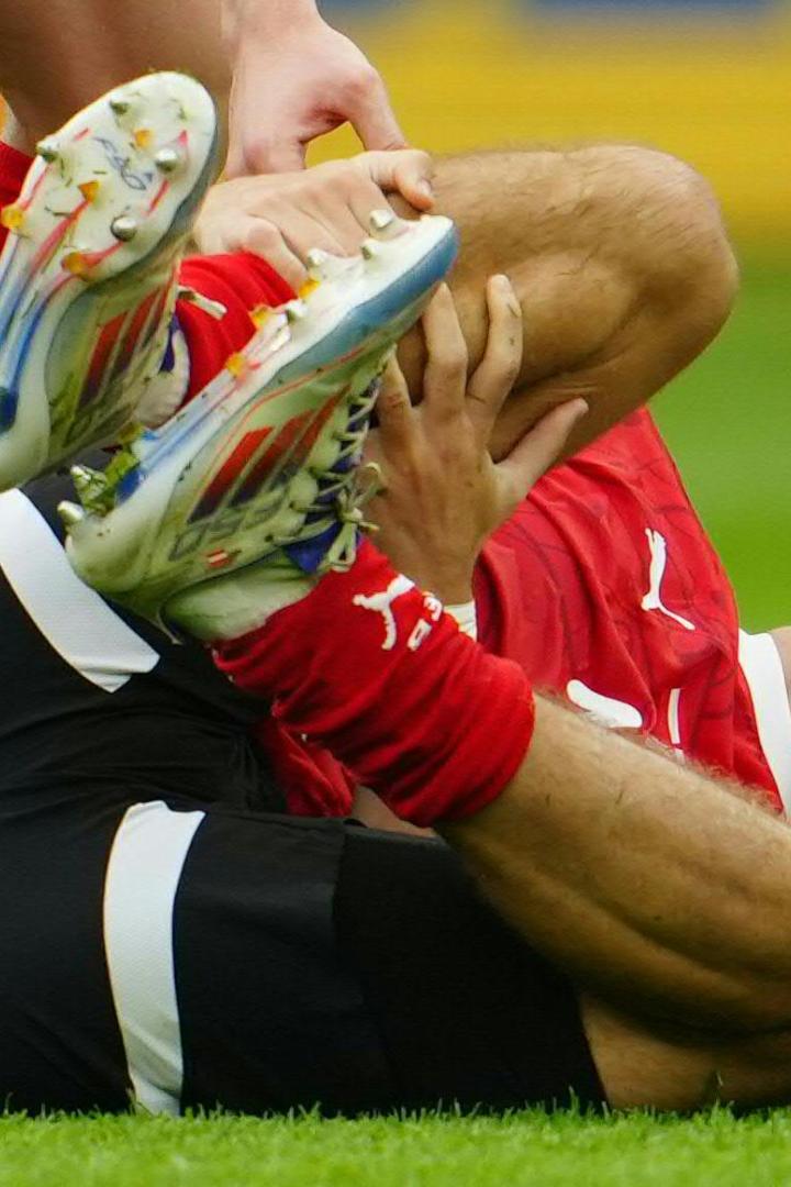Ein Fußballspieler in rotem Trikot liegt schmerzverzerrt auf dem Rasen und hält sich das verletzte Bein.
