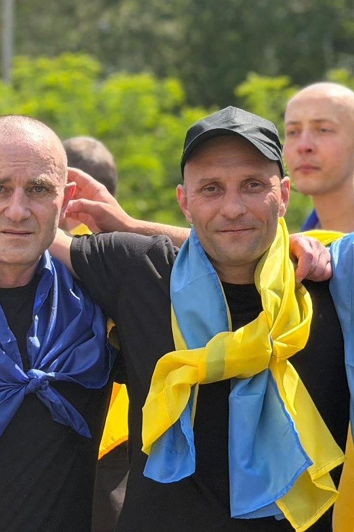 Männer, die in eine ukrainische Fahne gehüllt sind, legen sich die Arme um die Schultern und blicken in die Kamera.