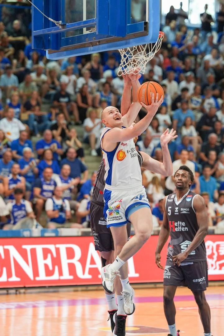 Ein Basketballspieler im weißen Trikot springt zum Korbleger, während ein Verteidiger im schwarzen Trikot versucht zu blocken.