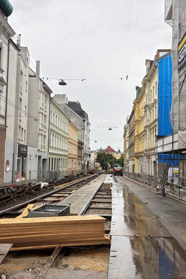 Straße mit Baustelle und offenen Straßenbahnschienen, umgeben von mehrstöckigen Häusern und Baugerüsten bei nassem Wetter.