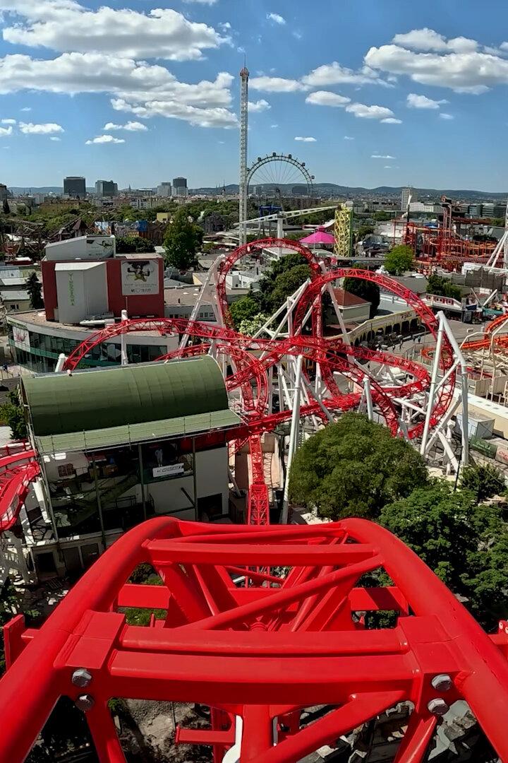 Blick von einer Achterbahn auf den Wiener Prater mit seinen Fahrgeschäften und dem Riesenrad unter einem sonnigen Himmel.