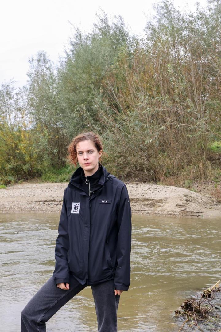 Eine Person steht am Ufer eines Flusses vor dichtem, herbstlichem Gebüsch und trägt eine schwarze Jacke mit WWF-Logo.