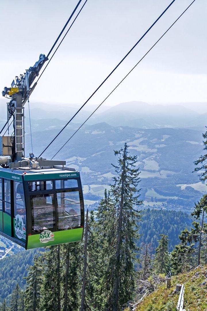 Eine grüne Seilbahngondel fährt über bewaldete Berghänge mit weitem Blick ins Tal und auf umliegende Berge.
