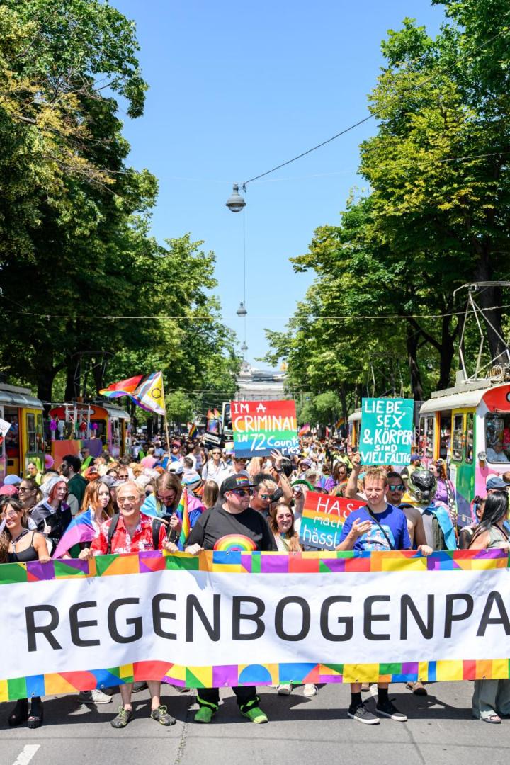 Viele Protestierende auf die Vienna ride Regenbogenparade halten einen großen banner. Im Hintergrund sind bunte Fahnen.