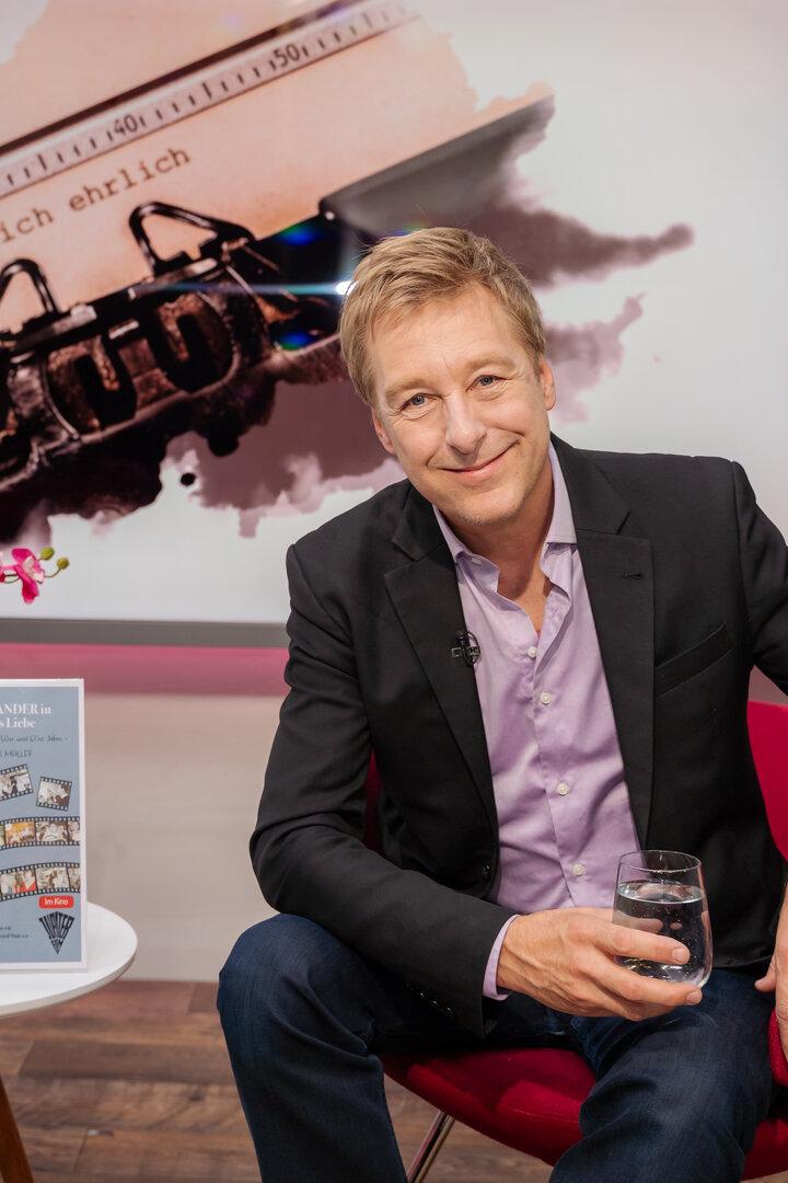 Max Müller sitzt in einem roten Sessel vor einem Plakat über Peter Alexander und hält ein Glas Wasser in der Hand.