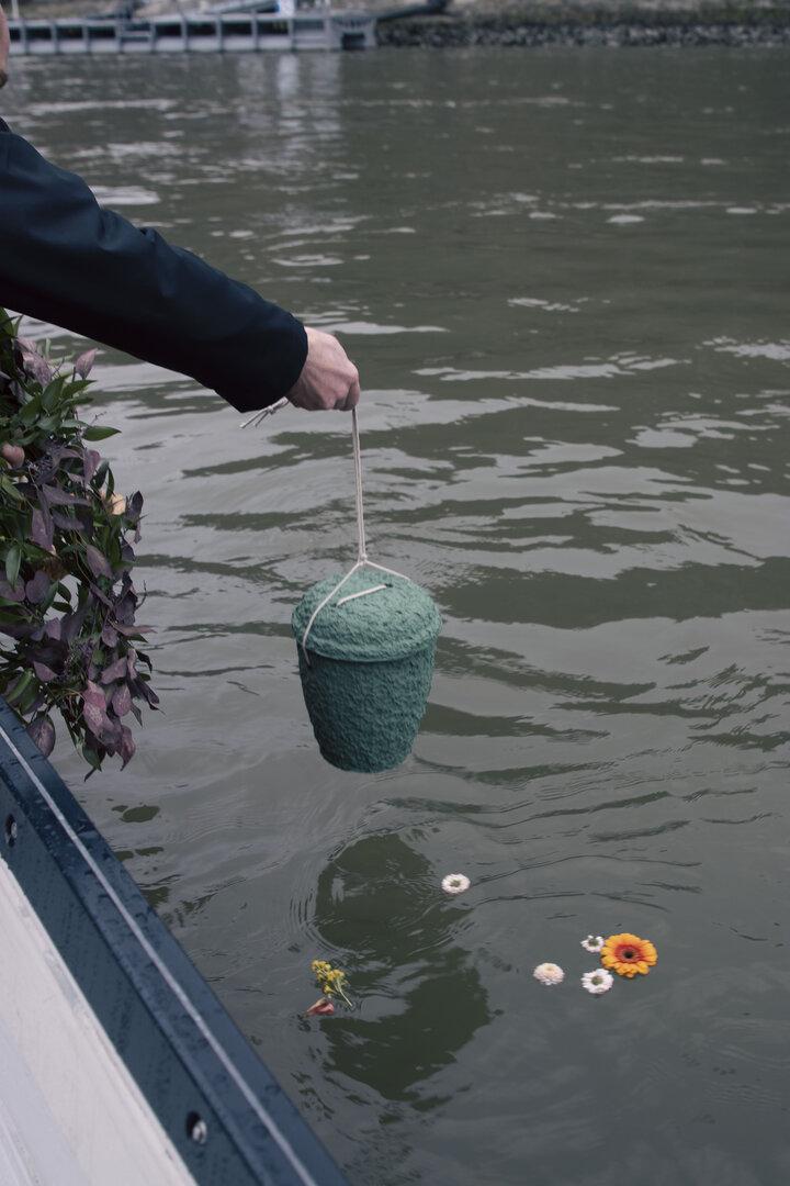 Eine Person hält eine grüne Urne über das Wasser, während Blumen auf der Wasseroberfläche treiben.