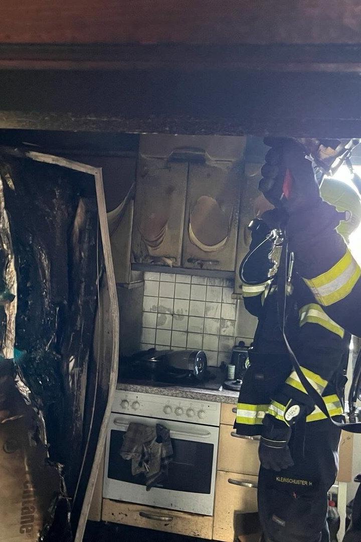 Zwei Feuerwehrleute in Schutzkleidung löschen einen Brand in einer stark beschädigten Küche mit verbrannten Geräten und Möbeln.