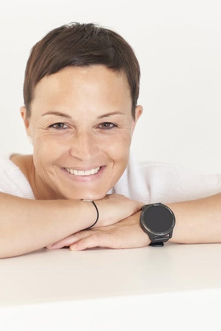 Frau mit kurzen braunen Haaren lächelt und stützt ihr Kinn auf verschränkte Arme, trägt eine schwarze Armbanduhr.