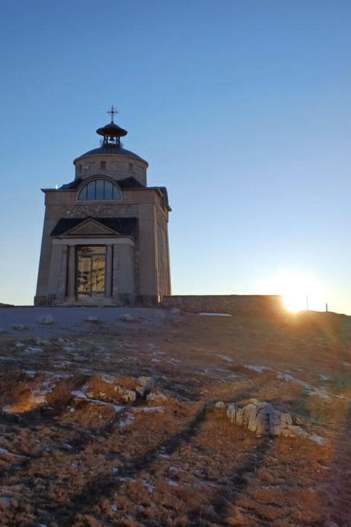 Kleine Kapelle auf einem Hügel bei Sonnenaufgang, mit klarem Himmel und spärlicher Vegetation im Vordergrund.